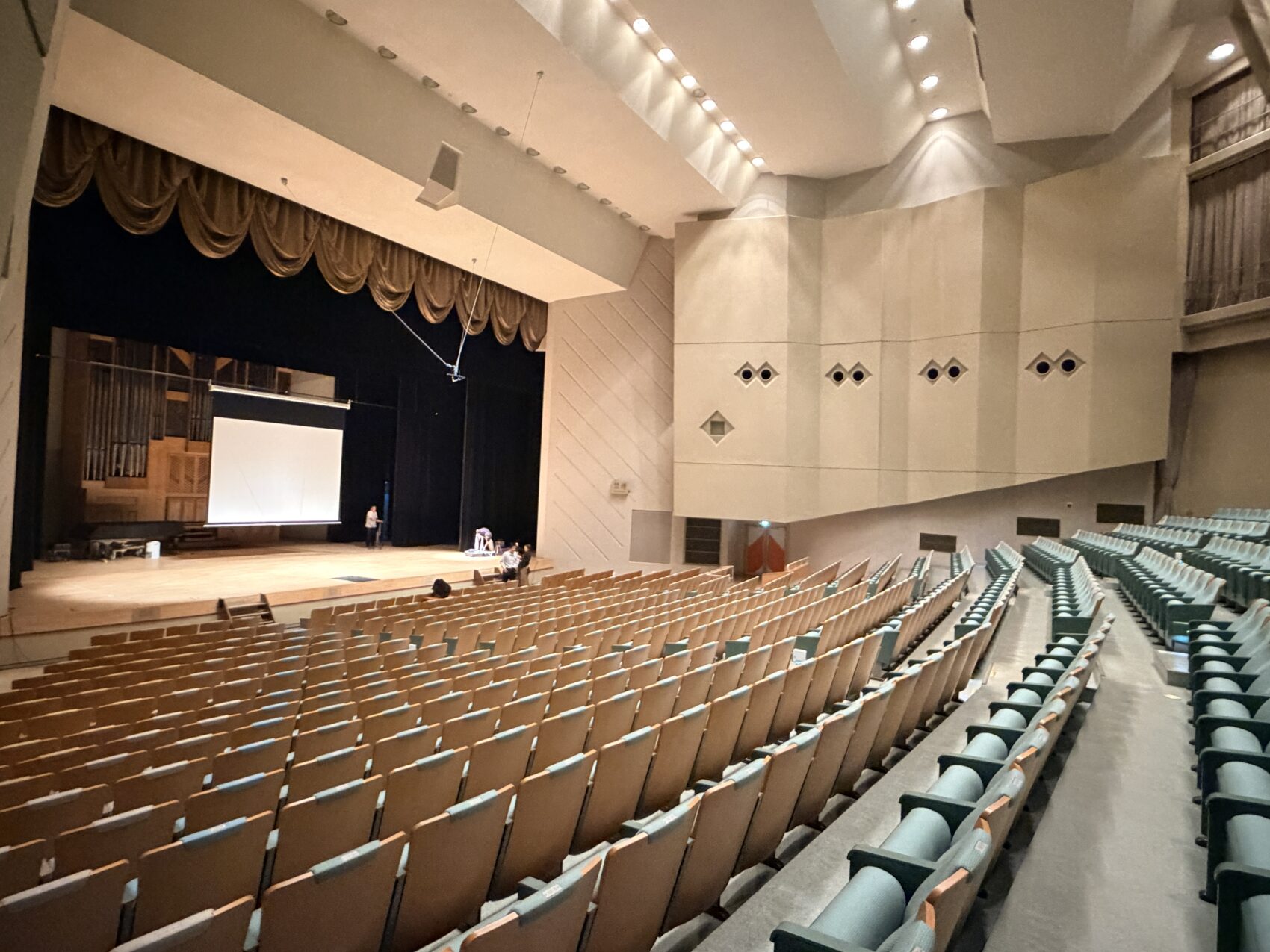 加美町バッハホールのステージと客席。講演の準備をする佐藤誠悦。
コンサートホールの客席からステージを見上げた広角写真。整然と並ぶ無人の座席と、奥のステージ上にある巨大なパイプオルガン、投影中の白いスクリーン、数人のスタッフが写っている。