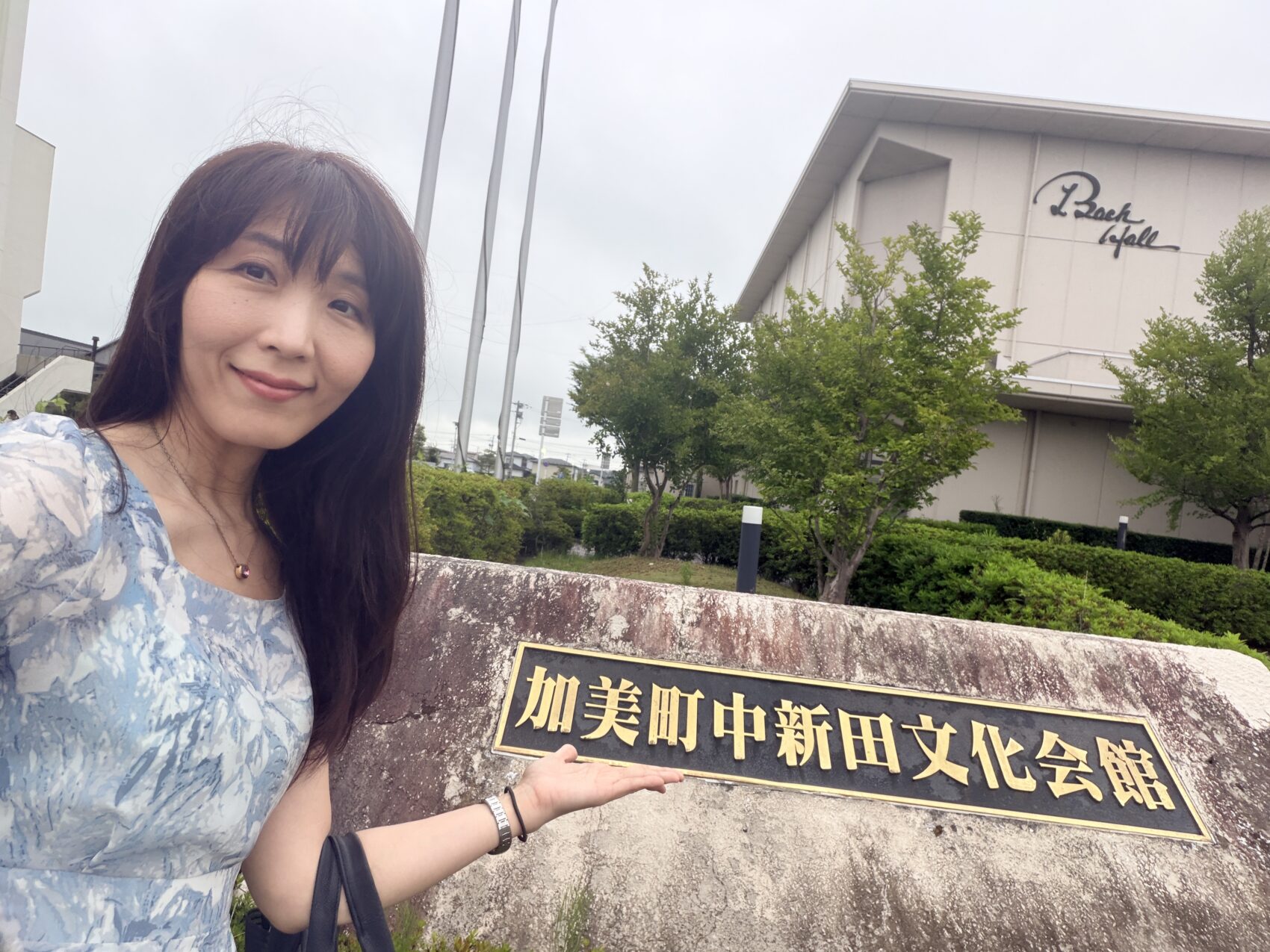 加美町中新田文化会館(中新田バッハホール)の入口にて。青い花柄ワンピース姿の女性が、「加美町中新田文化会館」と書かれた看板を紹介するセルフィー(自撮り)写真。奥にはホールの建物と「Bach Hall」のロゴが見える。