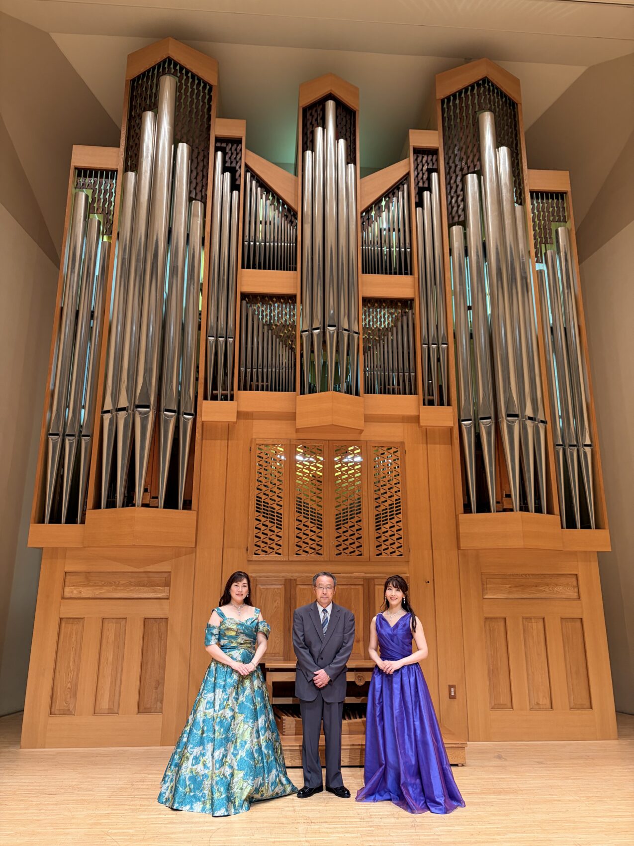 中新田バッハホールの巨大なパイプオルガンを背に、終演後のステージで記念撮影をする佐藤誠悦さん(中央スーツ)、氏家和歌子さん(左ブルー系ドレス)、藤波結花(右紫ドレス)。